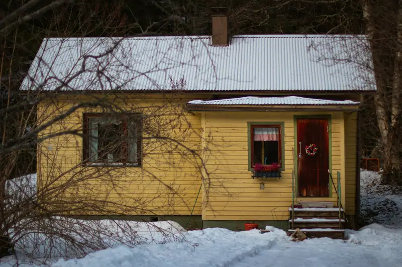 Keltainen puutalo talvella, ovessa havukranssi ja ikkunalaudalla punaisia kukkia.