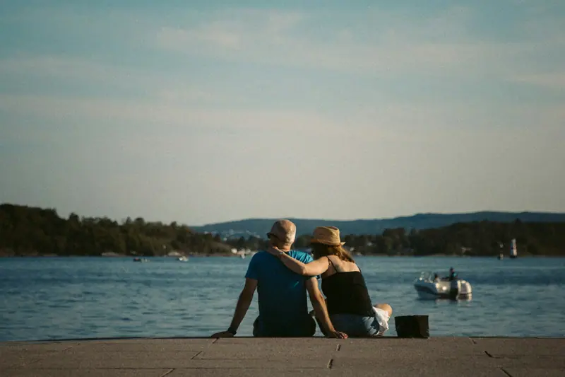 Kaksi ihmistä istuu vierekkäin laiturin reunalla ja katsoo merelle kesäiltana.