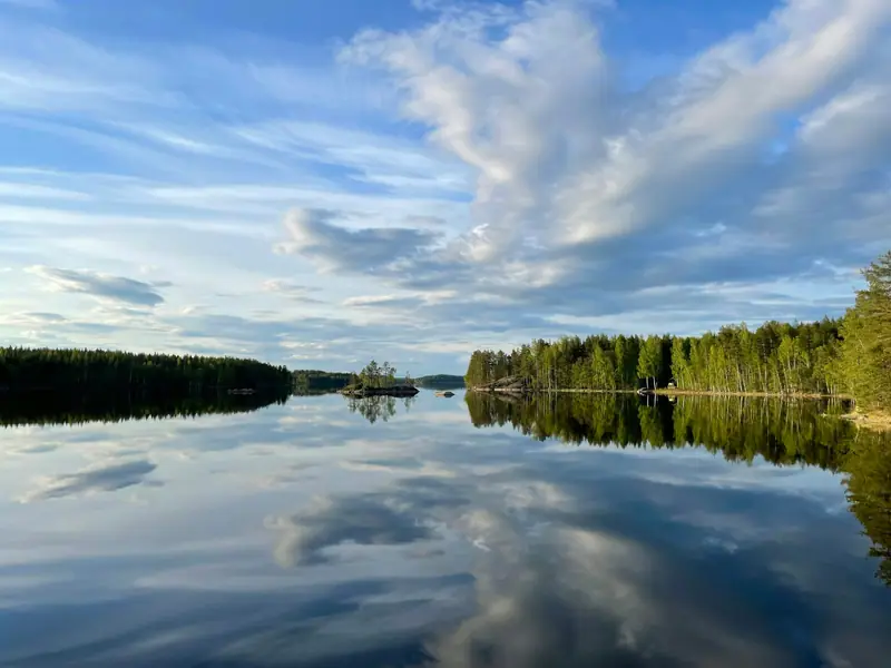 Tyyni järvi, jonka pintaan heijastuvat pilvet ja rantametsä.